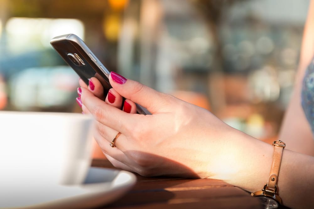 Hände halten ein Smartphone, im Hintergrund eine Kaffeetasse