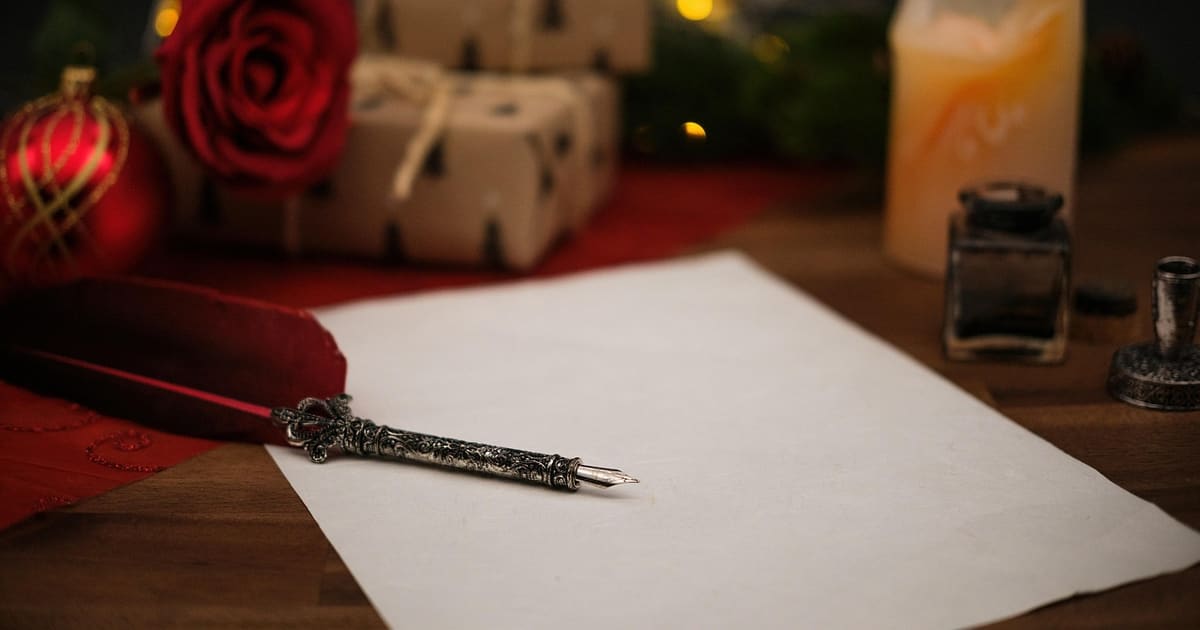 Weißes Briefpapier mit Federhalter auf einem Holztisch, dahinter Geschenke, eine rote Rose und eine Kerze